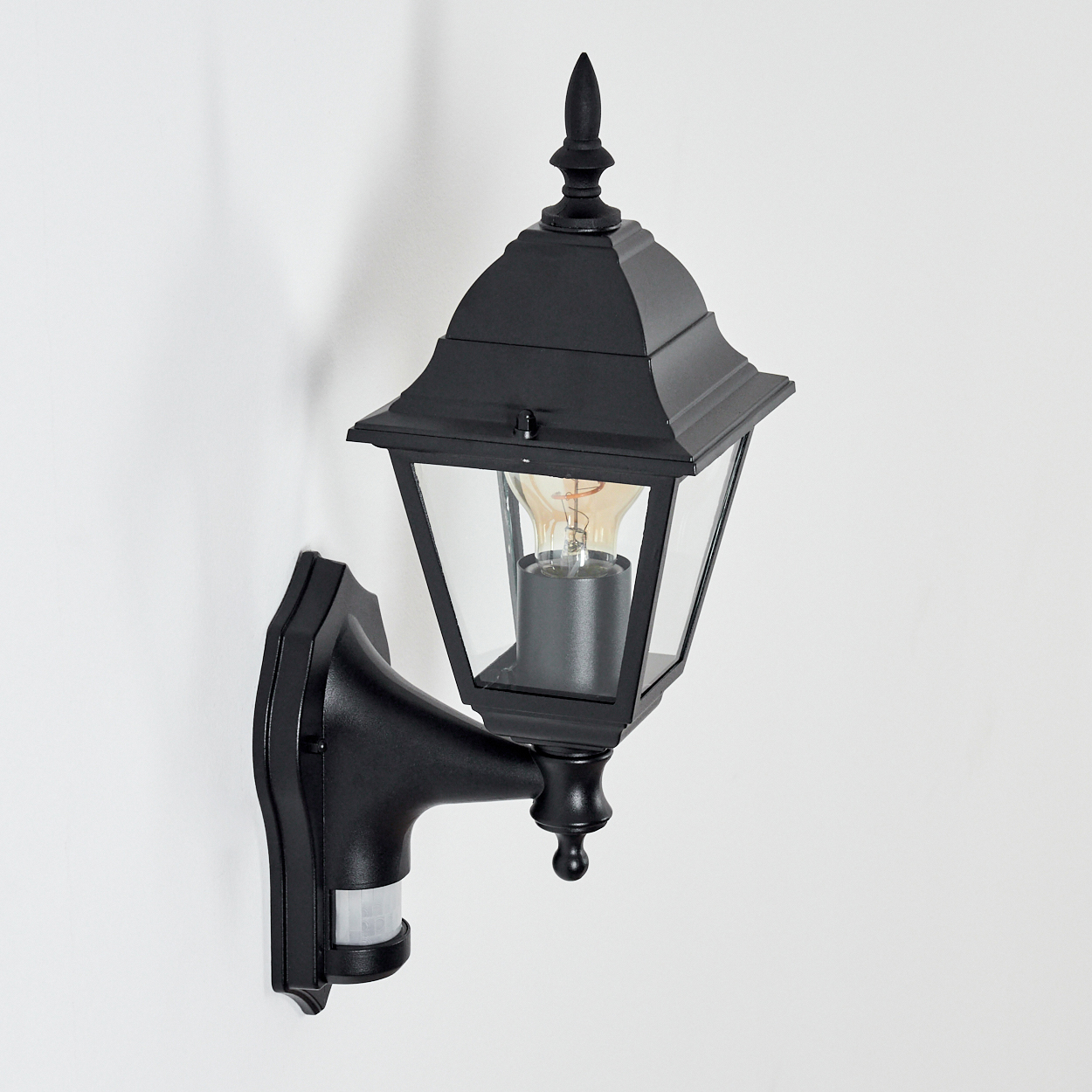 Eine schwarze Außenwandleuchte mit Glasgehäuse und sichtbarer Glühbirne. Sie ist an einer weißen Wand montiert.