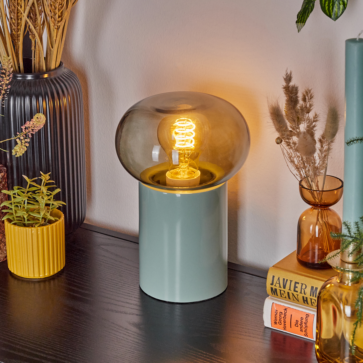 Eine moderne Lampe mit leuchtender Glühbirne steht auf einem schwarzen Tisch mit dekorativen Gegenständen wie Vasen und Büchern. Sanfte Beleuchtung.