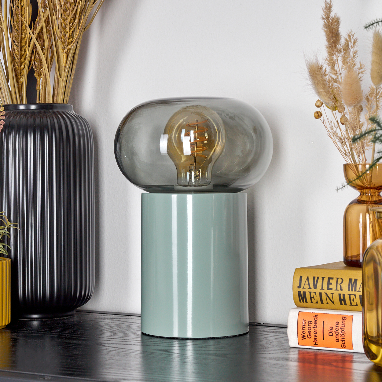 Eine moderne Lampe mit grünem Sockel und Glasschirm. Daneben Vasen mit Weizen und Bücher auf einem schwarzen Tisch.