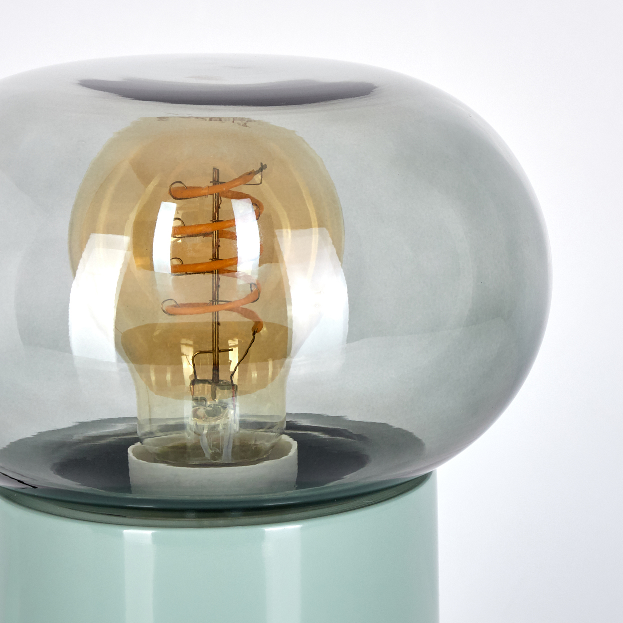 Eine moderne Lampe mit Glasschirm und sichtbarer Glühbirne. Der Sockel ist mintgrün, der Schirm grau und die Glühbirne orange.