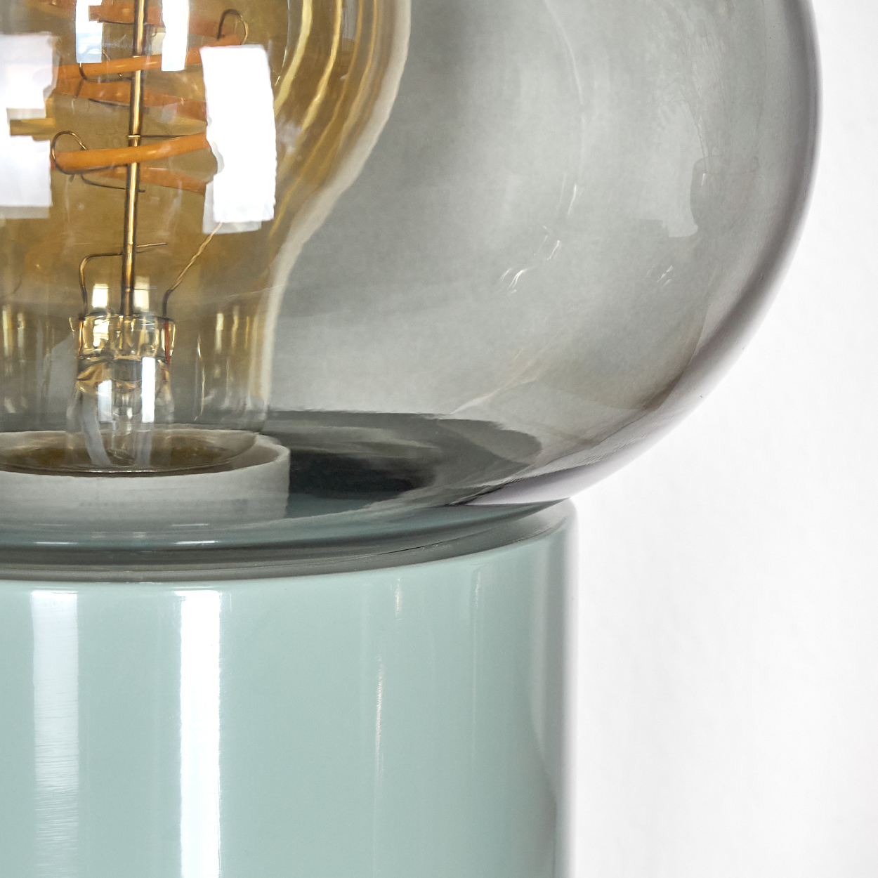 Nahaufnahme einer Leuchte. Sie hat einen blau-grünen Sockel, eine klare Glühbirne mit goldenem Glühfaden und einen rauchgrauen Glaskolben.