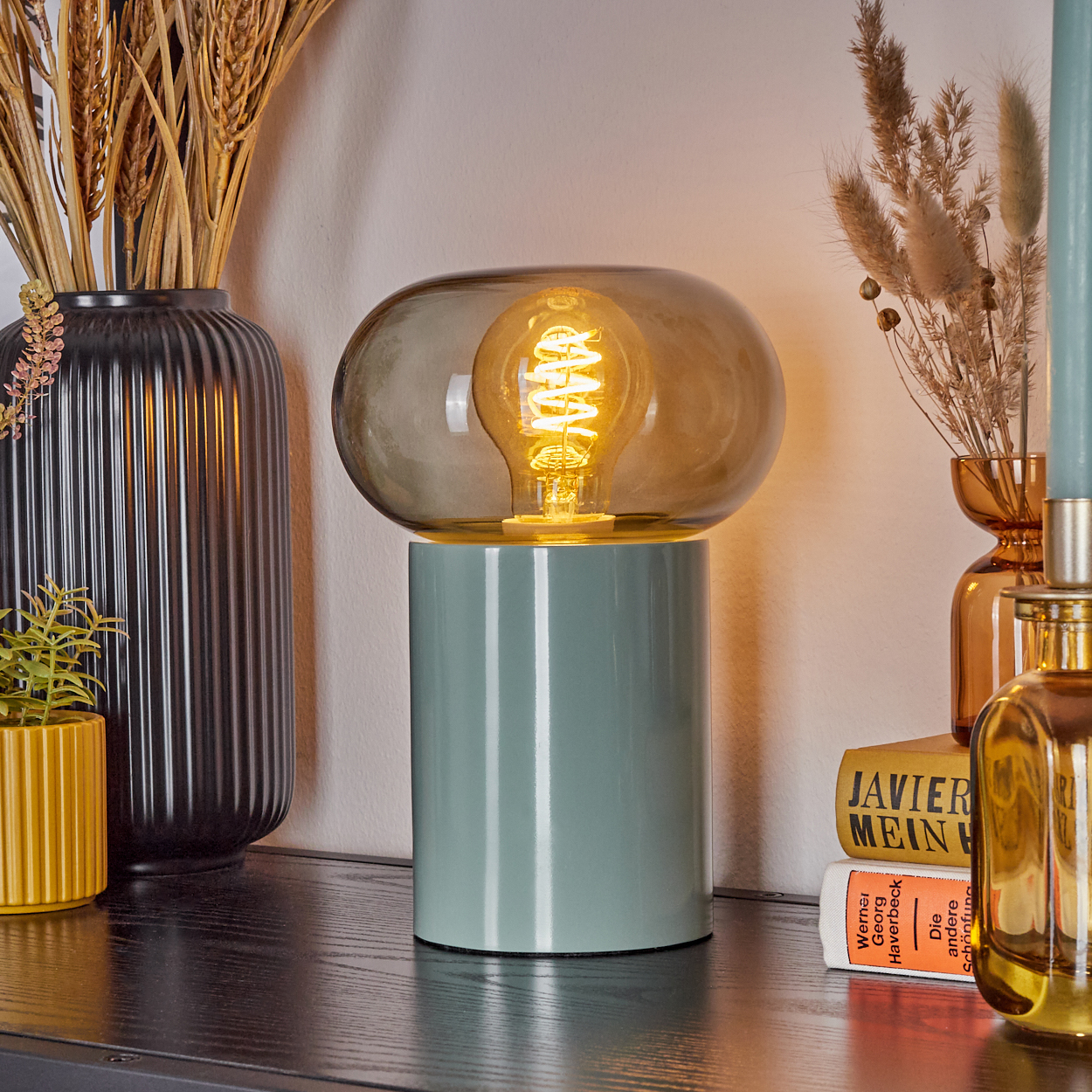 Eine moderne Lampe mit einer Glühbirne in einem Glasschirm steht auf einem Regal. Vasen, eine Pflanze und Bücher sind darum herum.