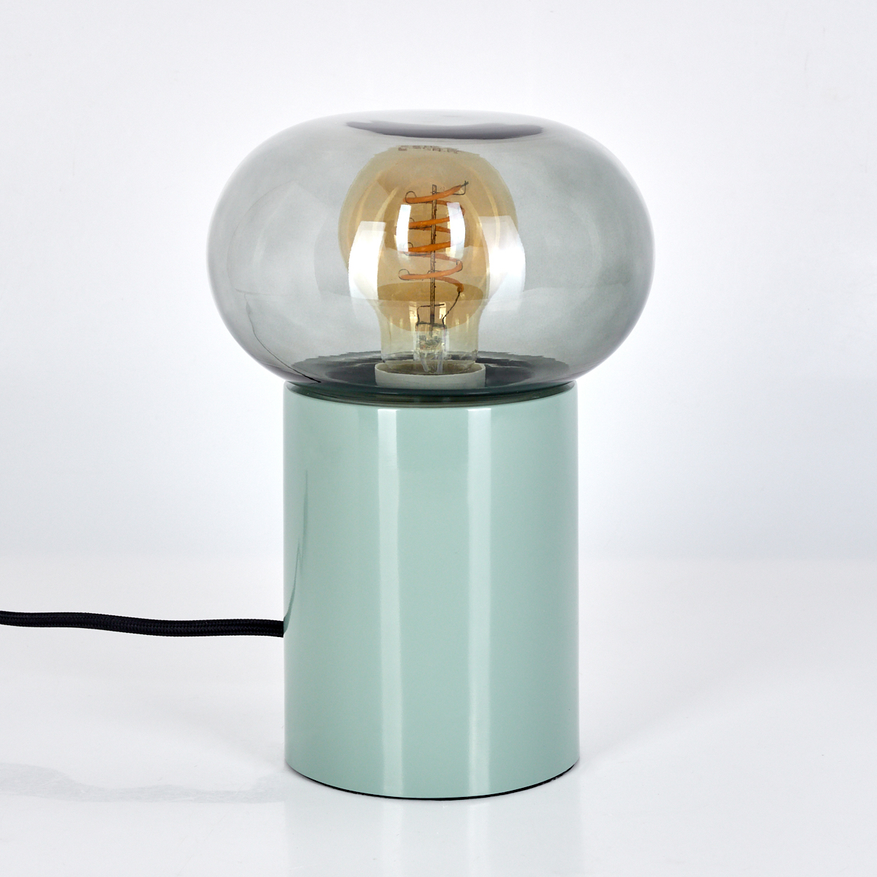 Eine moderne Lampe mit hellgrüner zylindrischer Basis, grauem Glasschirm und sichtbarer Glühbirne. Schwarzes Kabel.