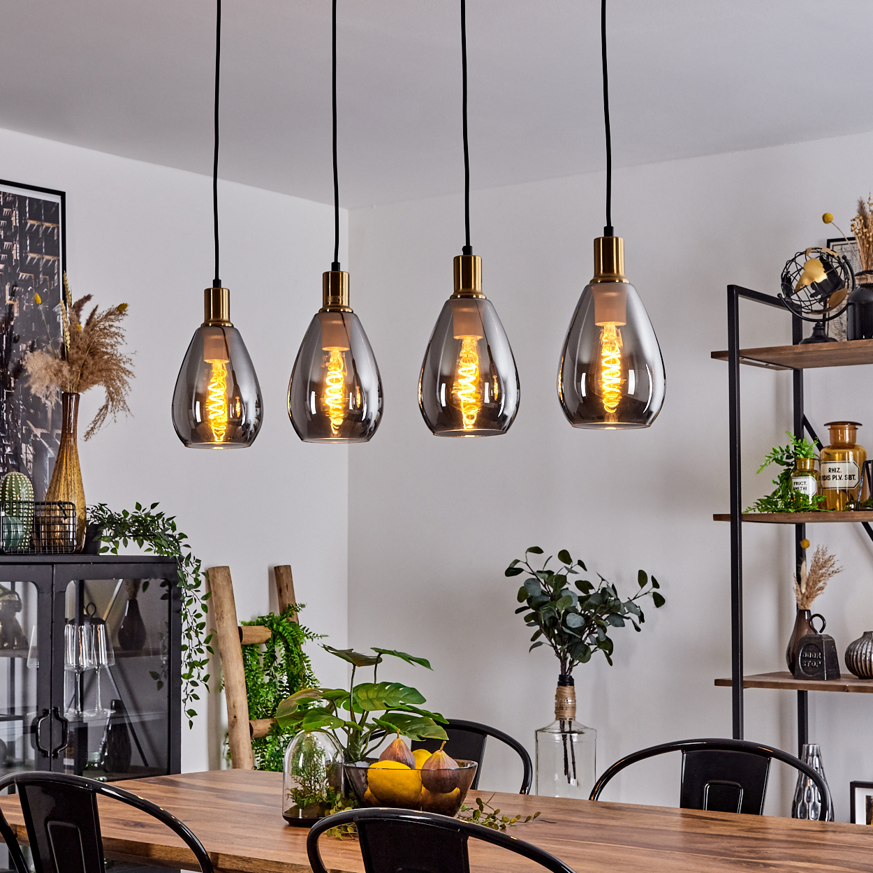 Vier hängende tropfenförmige Lampen mit goldenen Akzenten über einem Holztisch. Pflanzen und Dekoration umgeben den Tisch.