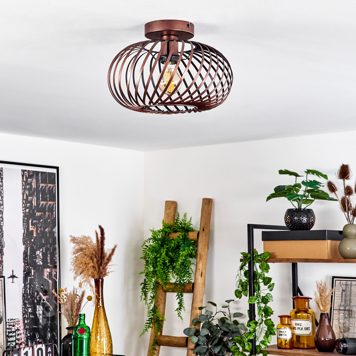 Über verschiedenen Dekorationsartikeln hängt eine bronzefarbene, käfigartige Deckenlampe. Eine Leiter, Pflanzen und Flaschen sind darunter angeordnet.