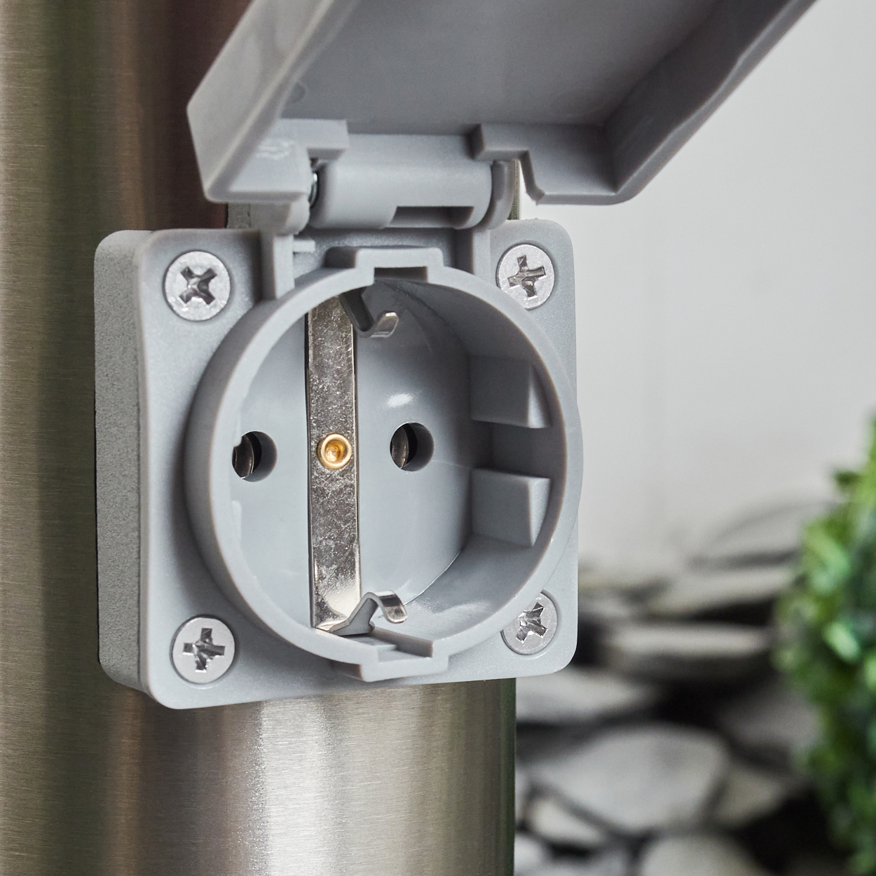 Nahaufnahme einer grauen Außensteckdose mit offenem Deckel, montiert an einem Metallpfosten. Schrauben sichern die Steckdose. Grüne Pflanzen sind im Hintergrund unscharf.