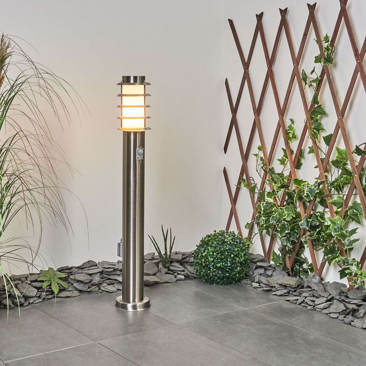 Eine moderne Außenlampe steht in einem Garten. Efeu klettert an einem Holzgitter. Grüne Pflanzen und graue Steine umgeben die Lampe.