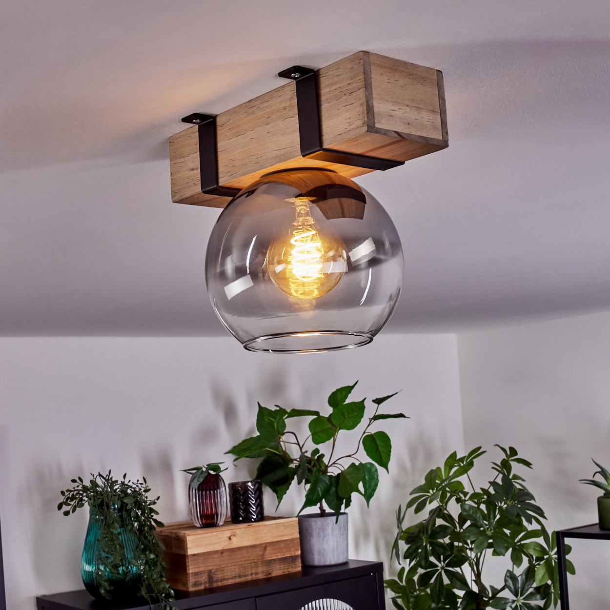 Eine Deckenleuchte mit Holzbalken und Glaskugel. Darunter Pflanzen auf einem Schrank.