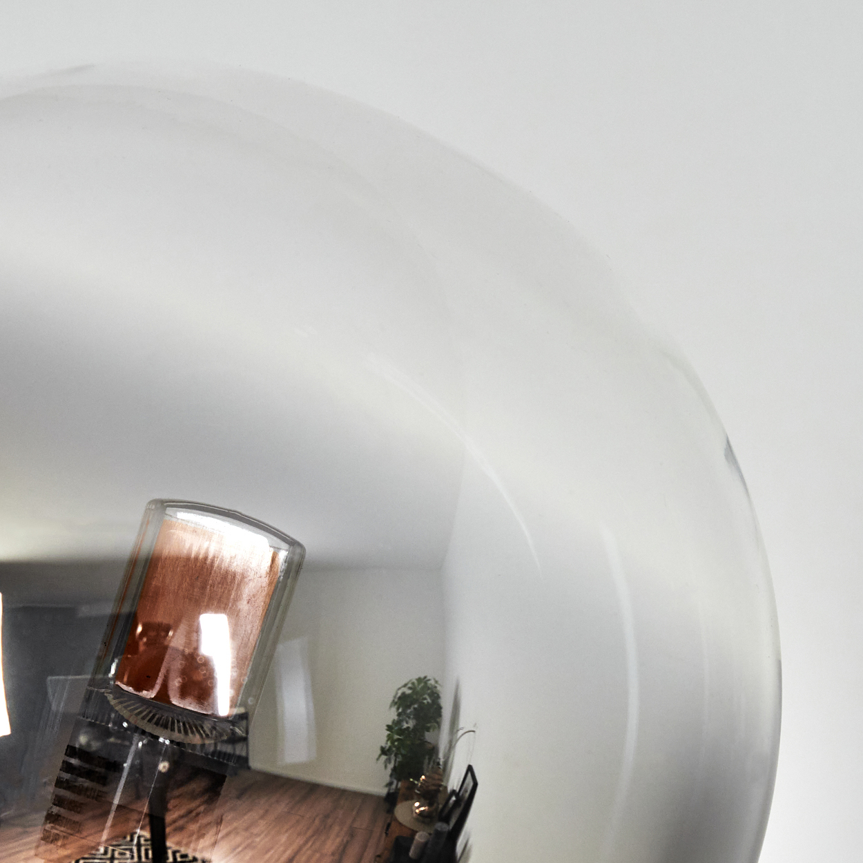 Eine reflektierende Oberfläche zeigt das Innere eines Raumes. Ein braunes rechteckiges Objekt ist sichtbar. Eine Pflanze und ein Holzboden sind zu sehen.