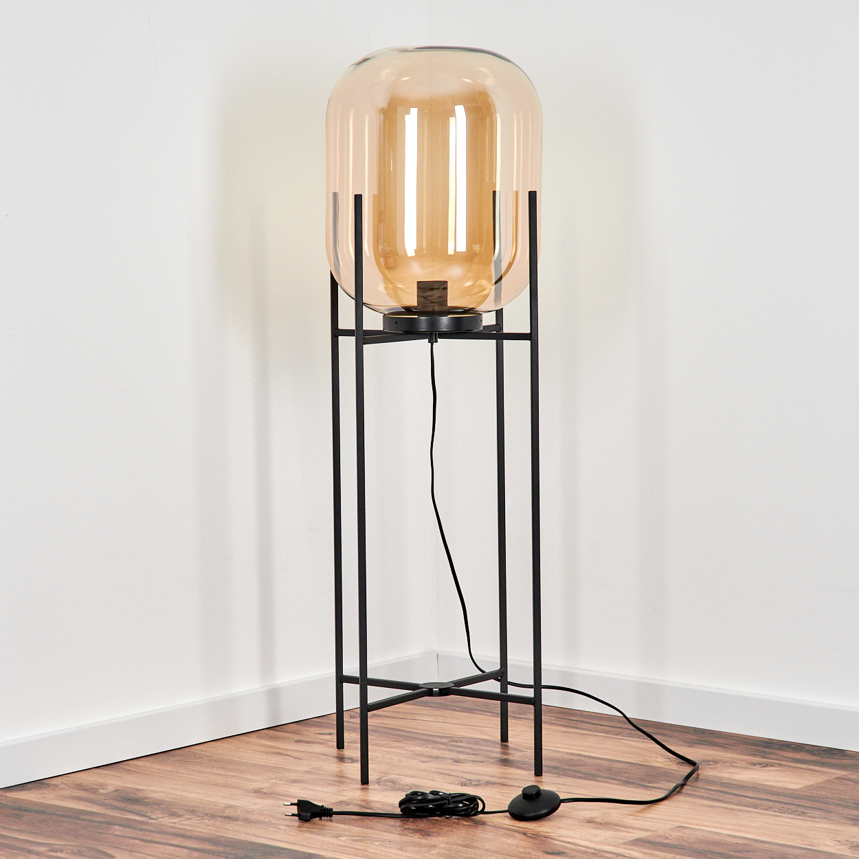 Eine moderne Stehlampe mit großem bernsteinfarbenem Glasschirm steht auf einem schwarzen Metallgestell. Sie steht auf einem Holzboden vor einer weißen Wand.