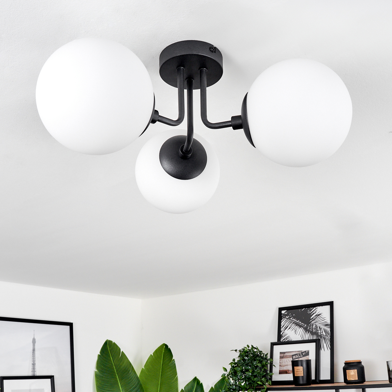 Eine Deckenlampe mit drei weißen Kugeln. Schwarze Metallarme und -halterung. Pflanzen und gerahmte Kunstwerke unten sichtbar.