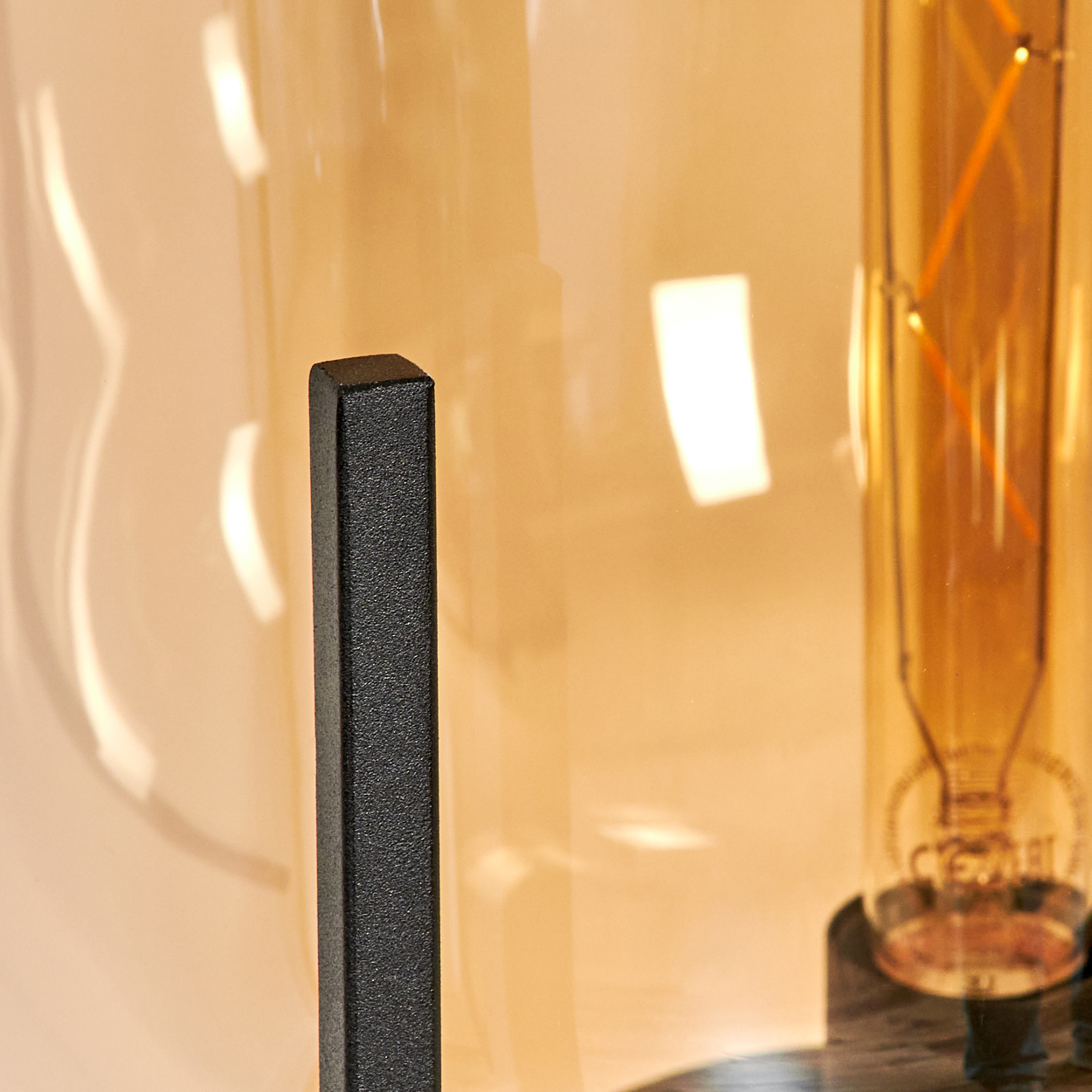 Nahaufnahme einer Lampe mit schwarzem Metallträger, Glasabdeckung und einer sichtbaren Glühbirne mit orangefarbenen Filamenten.