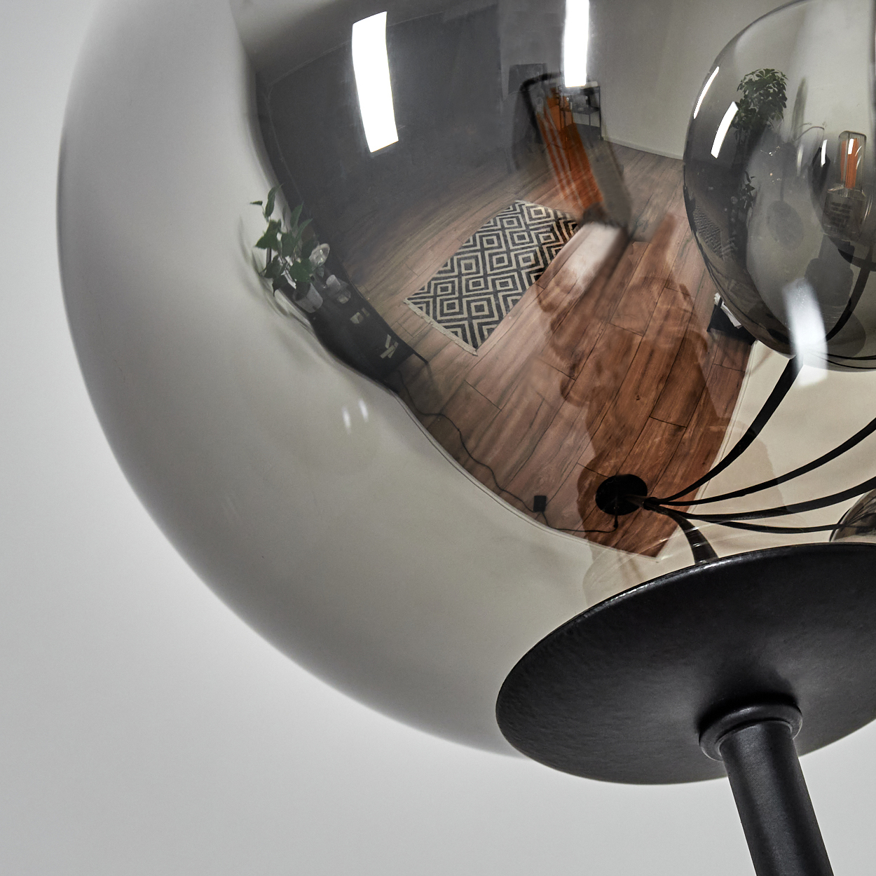 Eine große, kugelförmige Lampe spiegelt einen Raum mit Holzboden, einem gemusterten Teppich und einer Pflanze. Die Lampe hat einen schwarzen Fuß.