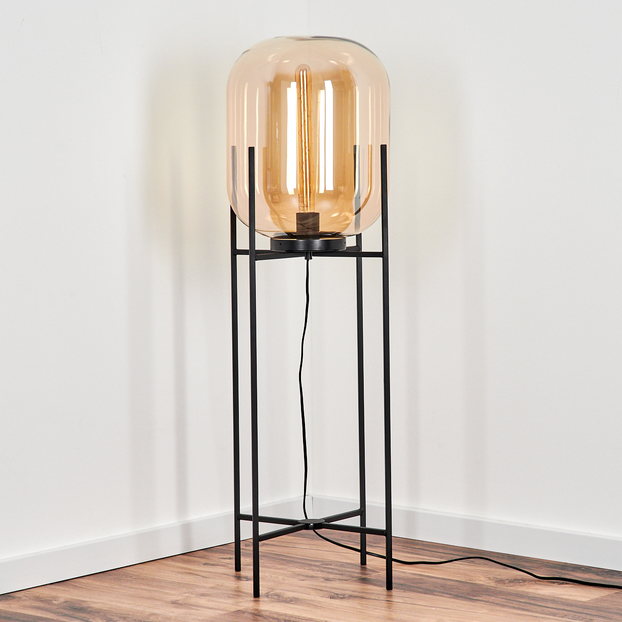 Eine hohe Stehlampe mit schwarzem Metallrahmen und bernsteinfarbenem Glasschirm. Sie steht auf einem Holzboden vor weißen Wänden.