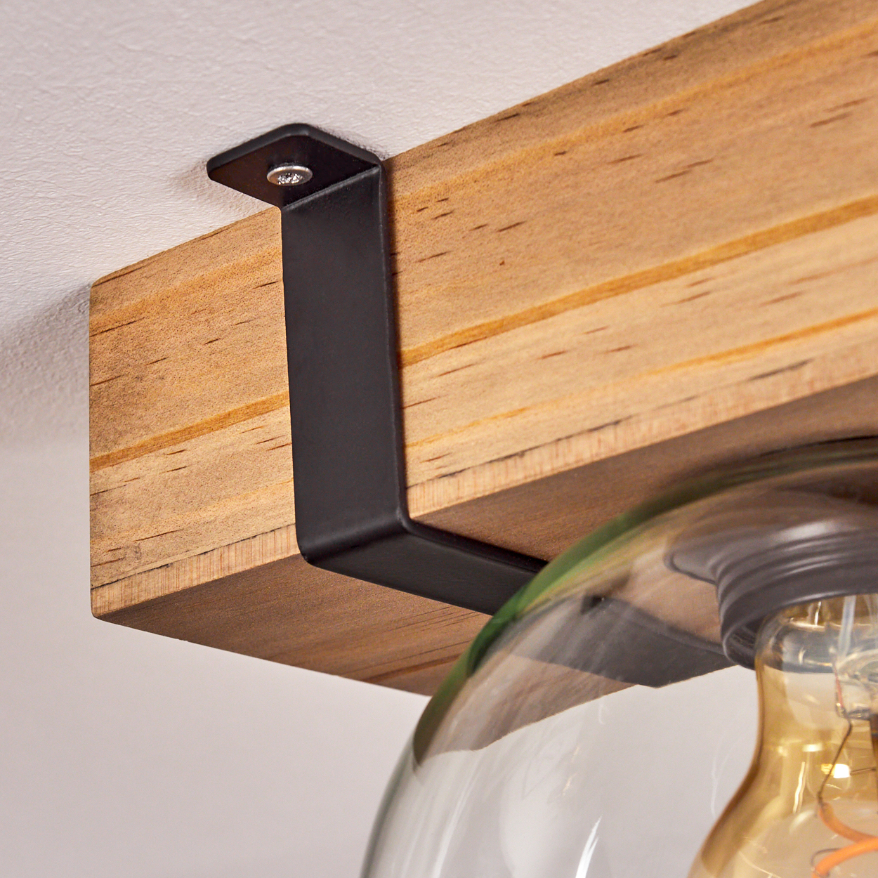 Nahaufnahme eines Holzbalkens, der mit einer schwarzen Metallhalterung an einer weißen Decke befestigt ist. Ein klarer Glasschirm und eine Glühbirne sind sichtbar.