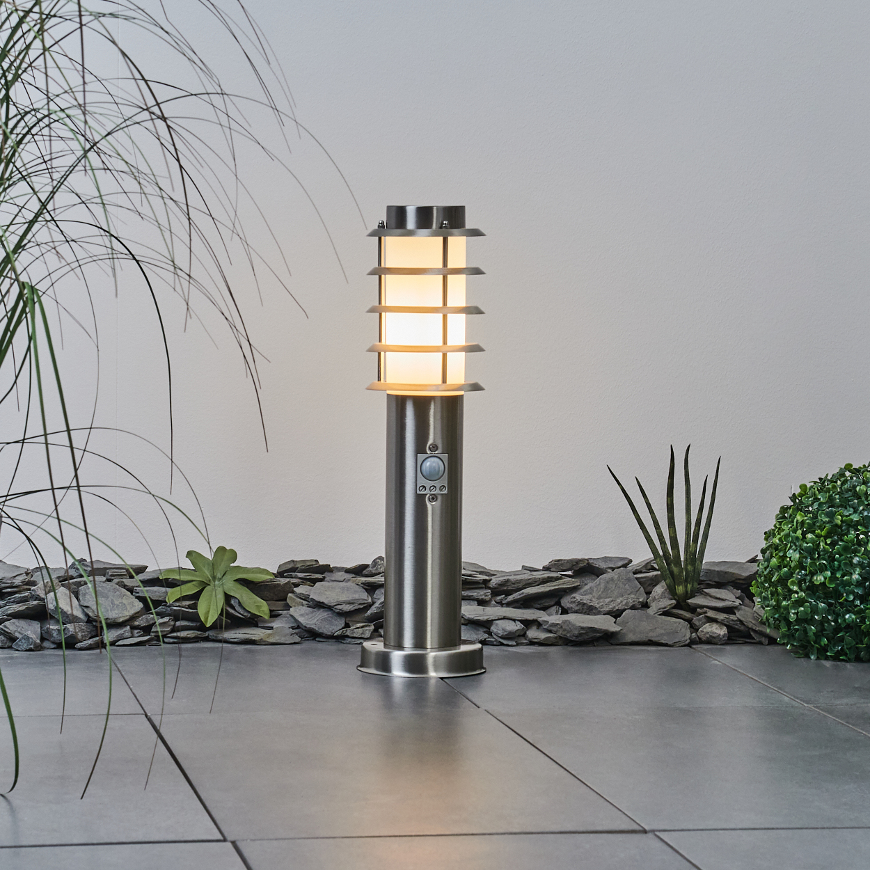 Eine beleuchtete Außenlampe mit Sensor steht auf einer gefliesten Oberfläche. Die Lampe ist von Pflanzen und dunklen Steinen umgeben.