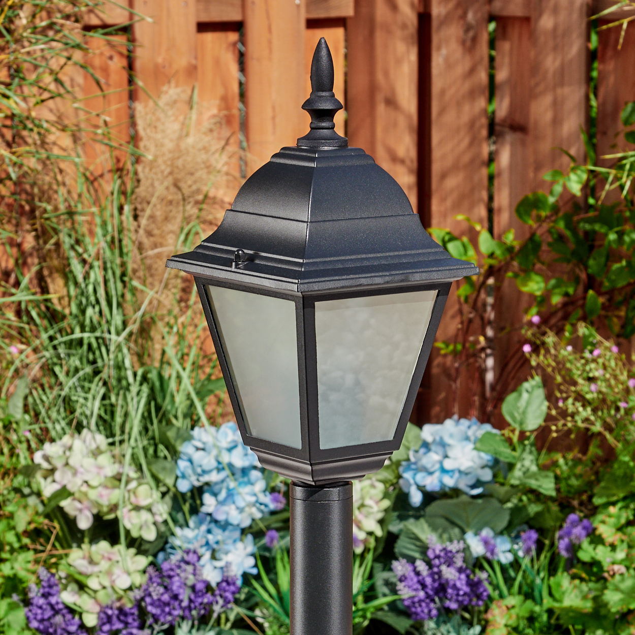 Schwarze Außenlampe auf einem Pfosten, umgeben von Blumen und Grün. Im Hintergrund befindet sich ein Holzzaun.