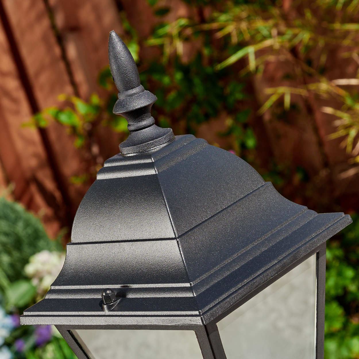 Nahaufnahme einer schwarzen Außenlampe mit dekorativer Spitze. Im Hintergrund ein Holzzaun und grünes Laub.