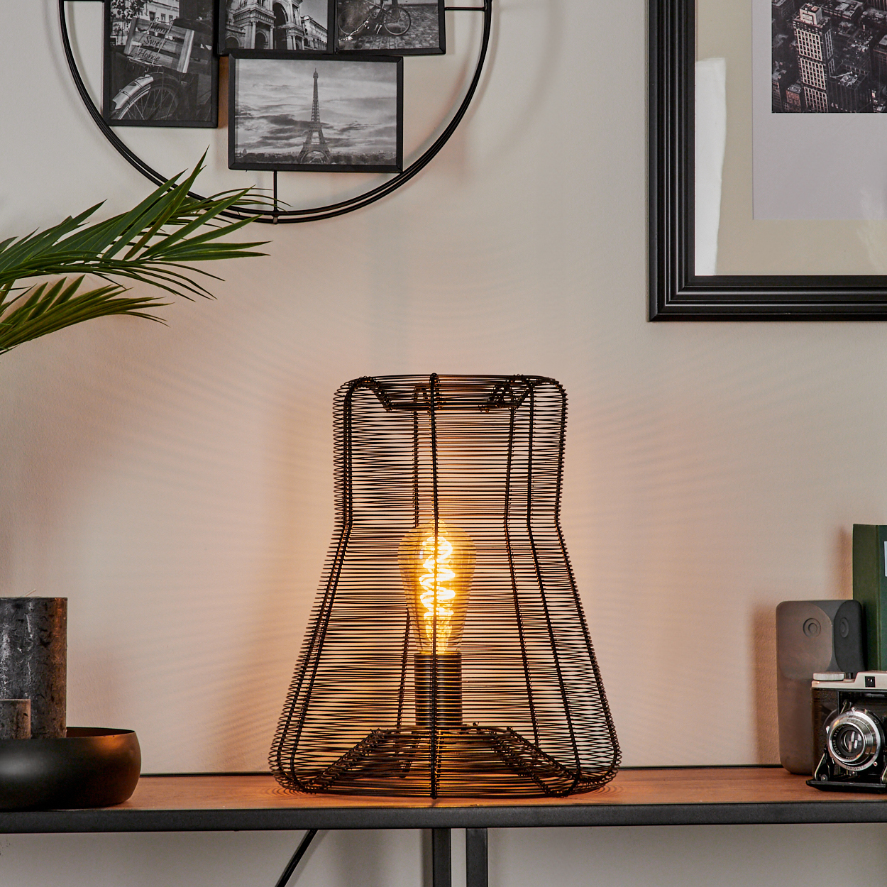 Eine schwarze Drahtlampe mit einer brennenden Glühbirne steht auf einer Holzoberfläche. Bilder und eine Pflanze sind im Hintergrund.
