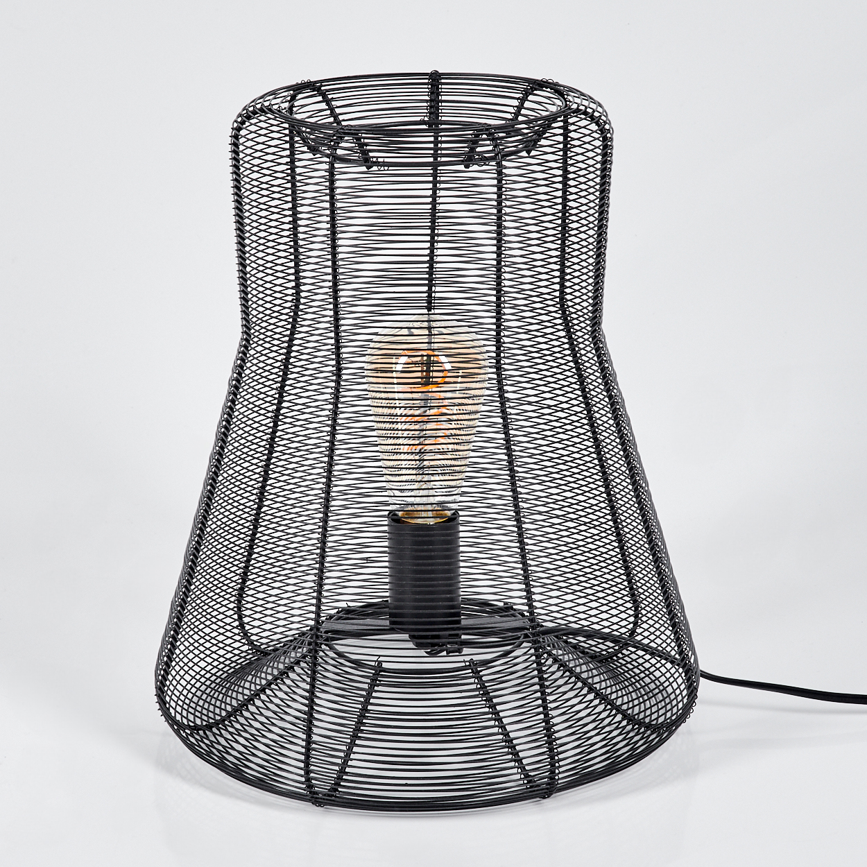 Schwarze Drahtrahmenlampe mit einer Glühbirne im Inneren. Sie steht auf einer weißen Oberfläche und hat ein schwarzes Netzkabel.