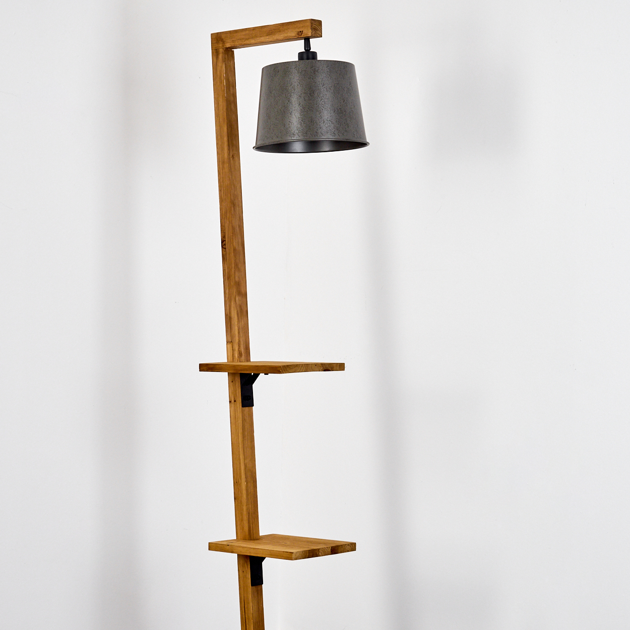 Eine Stehlampe aus Holz mit einem Lampenschirm aus Metall und zwei Regalen. Die Lampe steht vor einer weißen Wand.