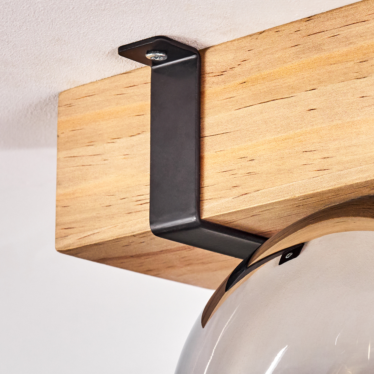 Ein Holzbalken wird von einer schwarzen Metallhalterung an einer weißen Decke gehalten. Darunter hängt eine Glaslampe.