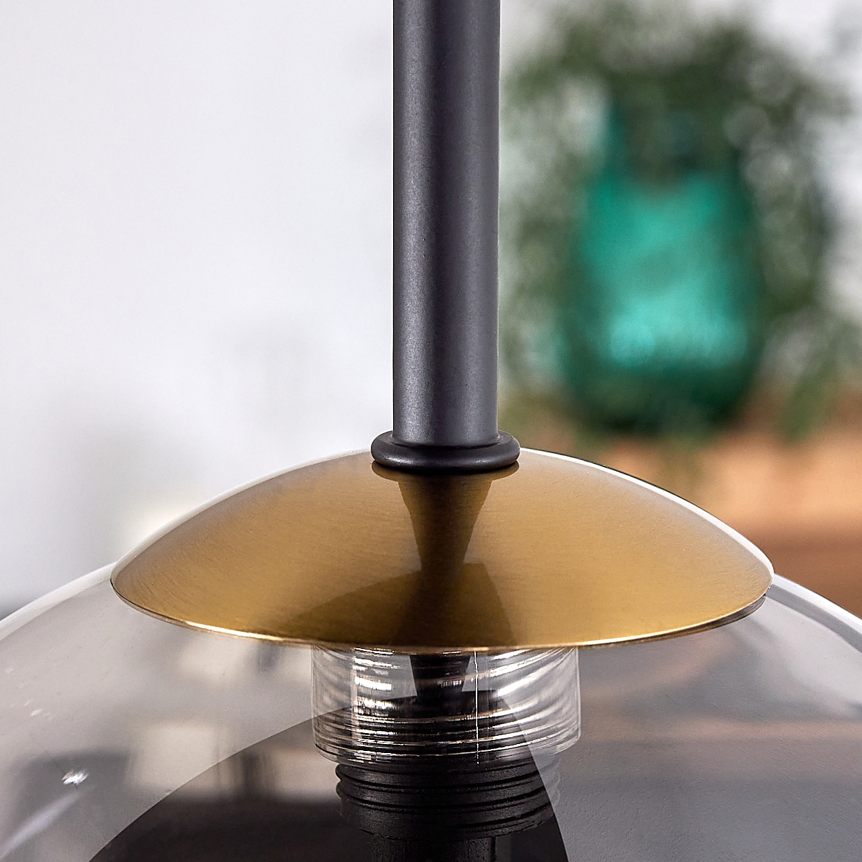 Nahaufnahme einer modernen Lampe. Sie hat einen schwarzen Stiel und einen goldfarbenen Schirm. Darunter ein Glasschirm. Im Hintergrund eine grüne Vase.