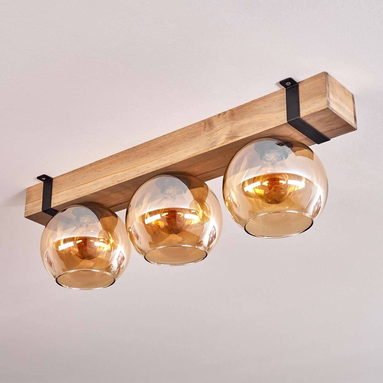 Eine Deckenleuchte mit drei bernsteinfarbenen Glaskugeln, die an einem Holzbalken hängen und mit schwarzen Metallhalterungen befestigt sind.