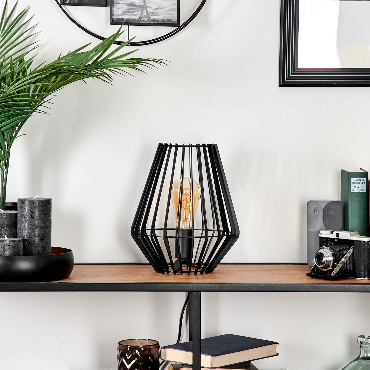 Schwarze Lampe mit sichtbarer Glühbirne auf einem Holzregal mit anderen Gegenständen. Ein Bild und eine Pflanze befinden sich im Hintergrund.