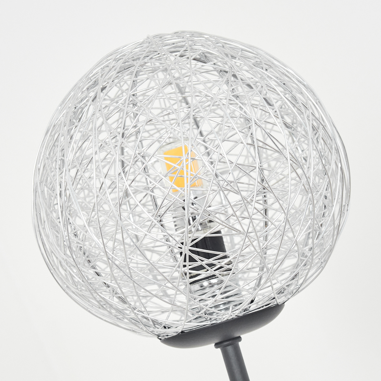 Eine dekorative Lampe mit kugelförmigem Drahtgestell. Im Inneren leuchtet eine Glühbirne. Der Sockel und der Stiel sind dunkel. Der Hintergrund ist weiß.