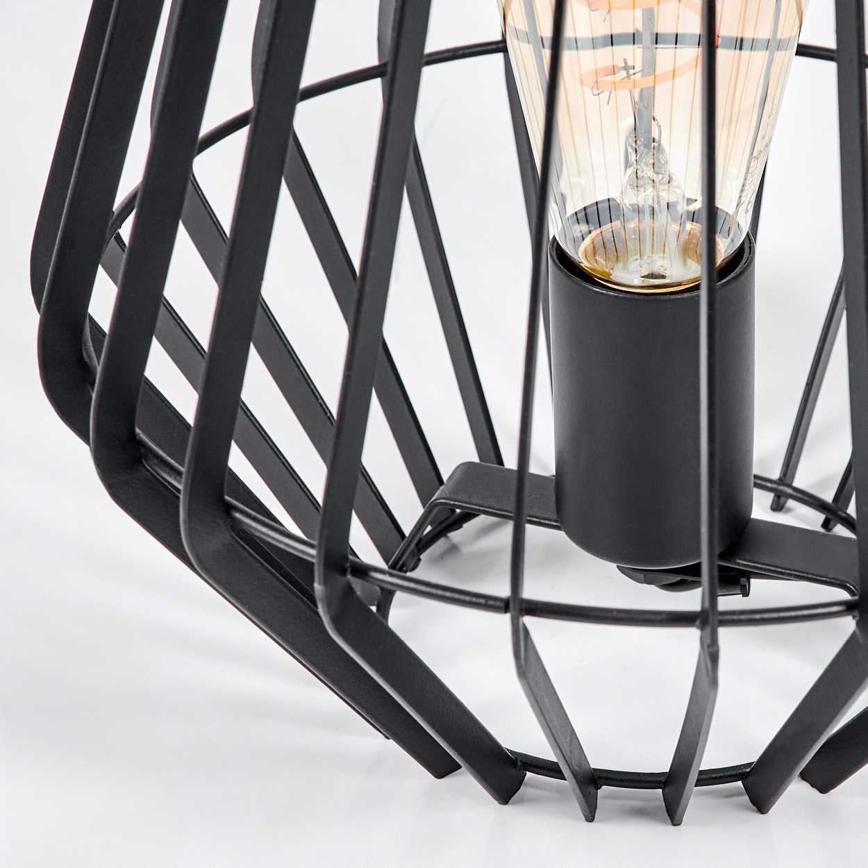 Eine schwarze Metallkäfiglampe mit einer Glühbirne. Die Lampe steht auf einer weißen Oberfläche. Die Glühbirne ist durch den Käfig sichtbar.