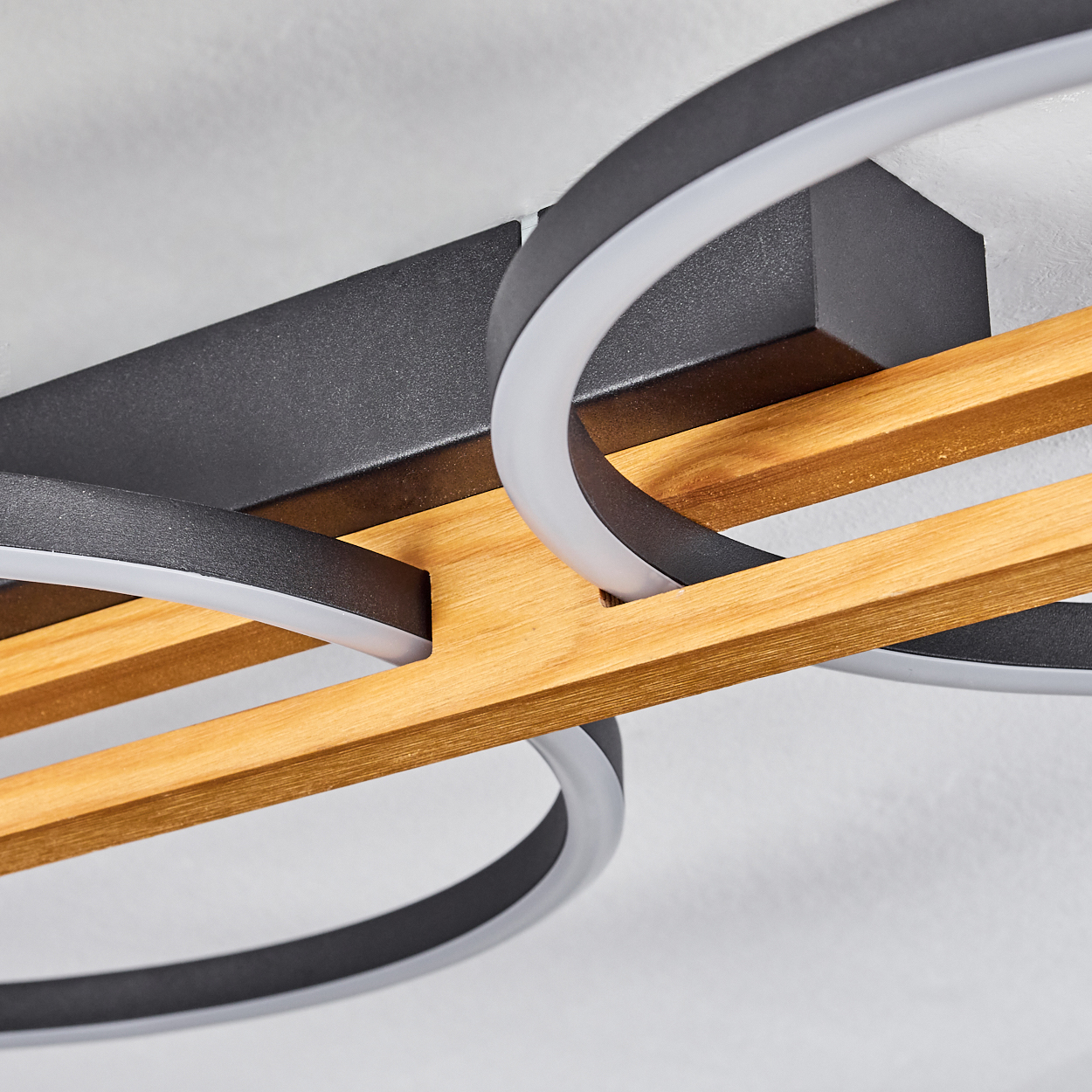 Moderne Deckenleuchte mit schwarzen Metallringen, Holzbalken und weißer Innenbeleuchtung, vor weißer Decke.