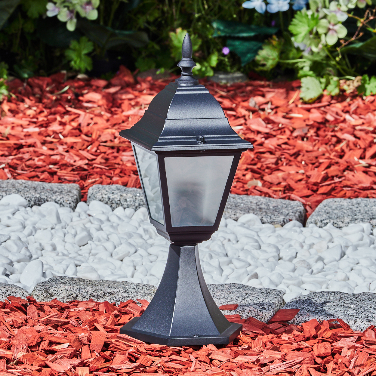 Eine schwarze Außenlampe steht auf roter Mulche, mit einem Hintergrund aus weißen Steinen, grauen Pflastersteinen und grünem Laub.
