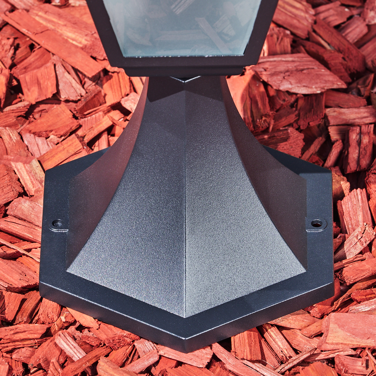 Ein schwarzer Außenlampensockel auf roter Mulch. Die Lampe hat ein geometrisches Design. Die Mulch besteht aus vielen Holzschnitzeln.