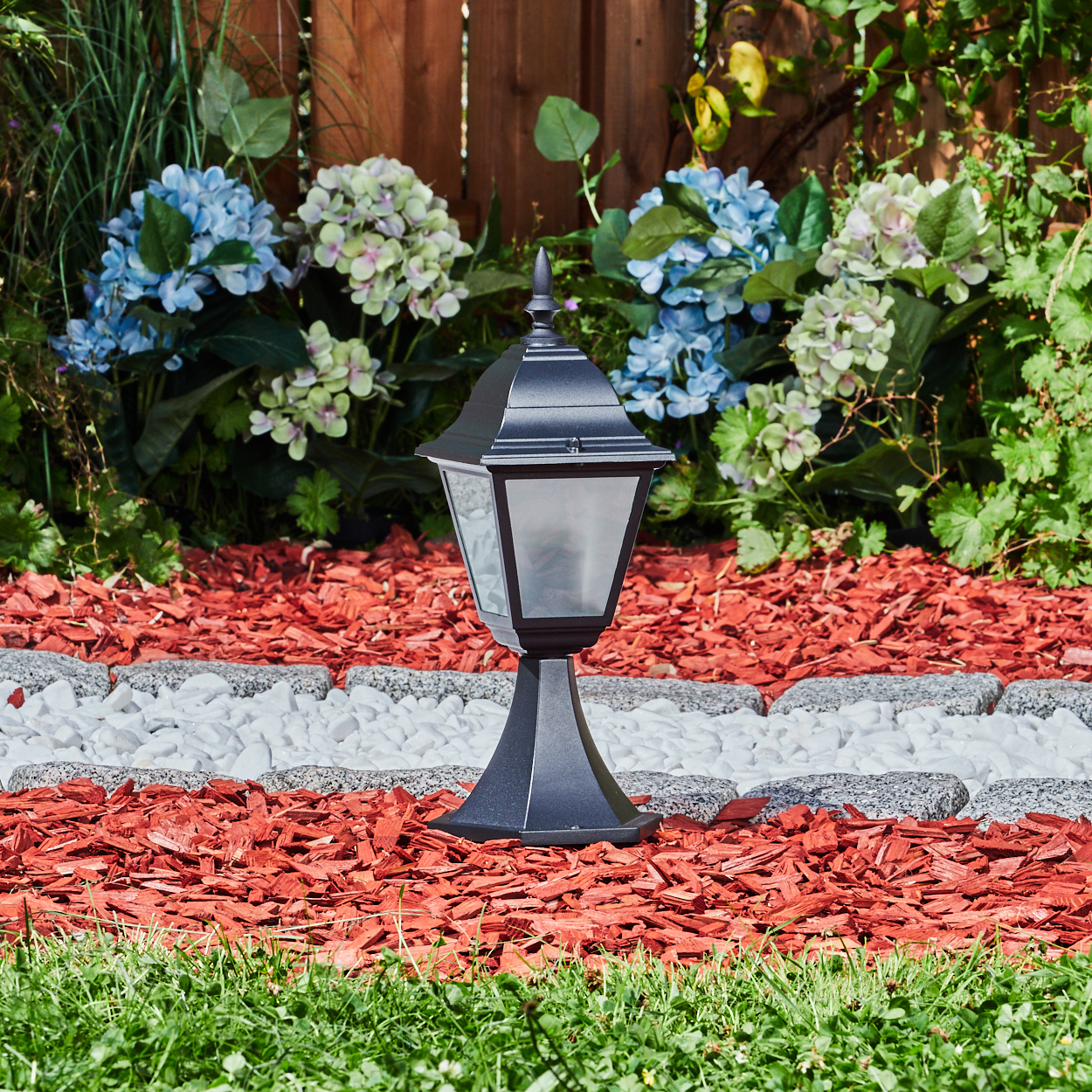 Schwarze Lampe im Garten. Rote Mulch, weiße Steine und grünes Gras. Blaue und weiße Blumen im Hintergrund. Holzzaun.