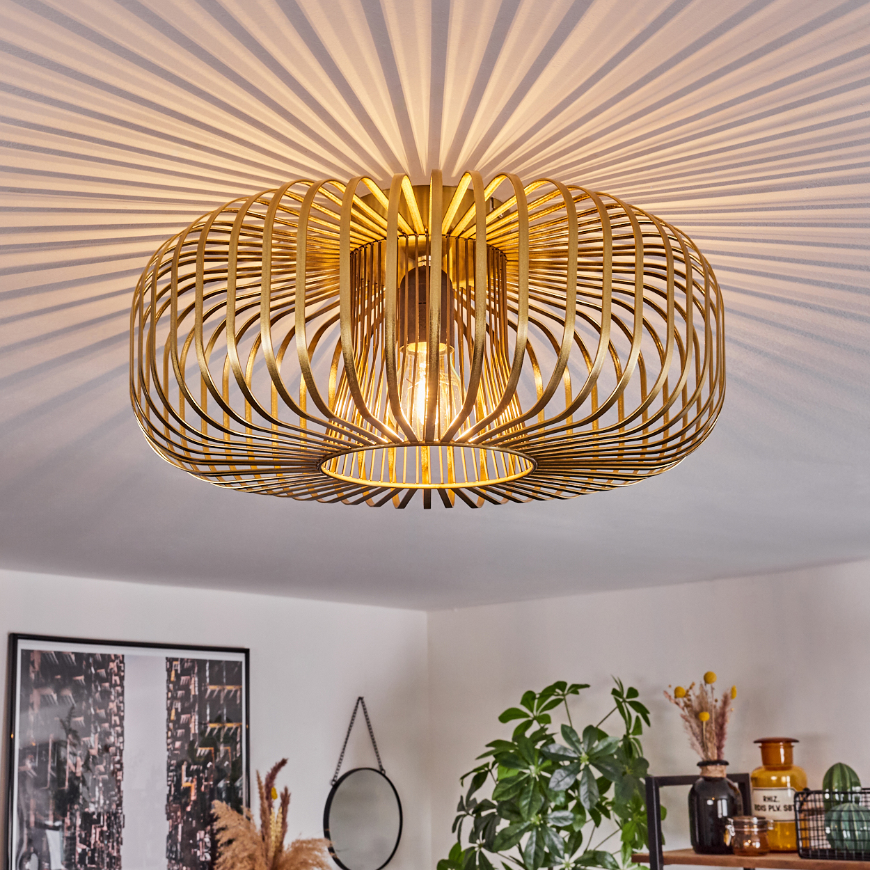 Eine goldene Deckenleuchte mit filigraner Metallarbeit, die Schatten auf die Decke wirft. Darunter ist ein Raum mit Pflanzen und Dekoration zu sehen.