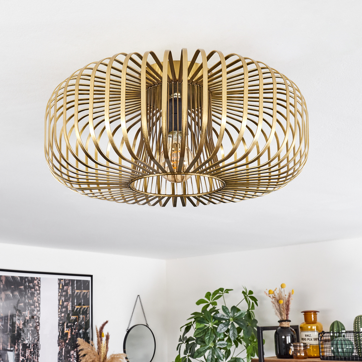 Eine goldene, käfigartige Deckenlampe ist zentriert. Der Hintergrund zeigt eine weiße Decke und ein dekoriertes Zimmer mit Pflanzen und einem Spiegel.