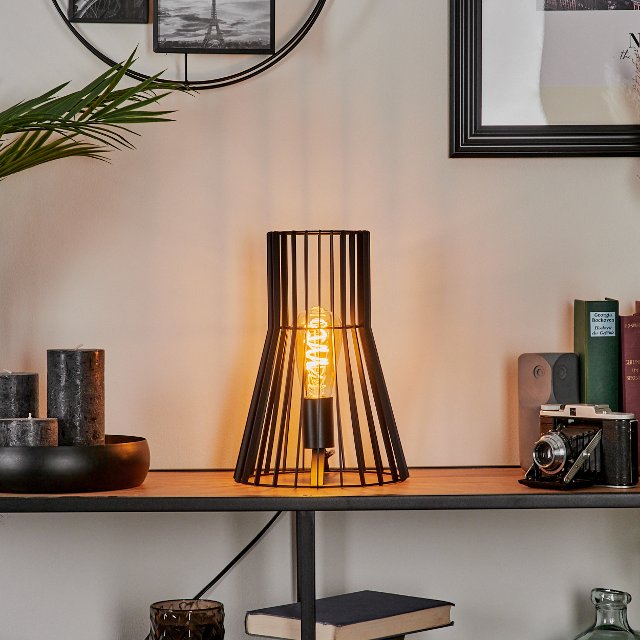 Eine schwarze Metalllampe mit sichtbarer Glühbirne steht auf einem Regal mit anderen dekorativen Gegenständen wie Kerzen und Büchern.