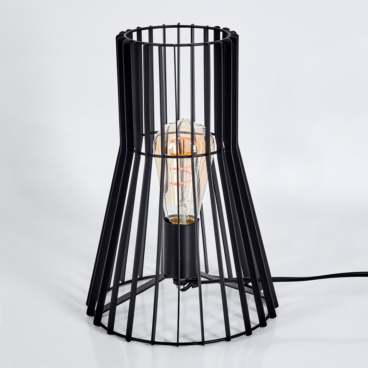 Eine schwarze Käfiglampe mit einer glühenden Edison-Glühbirne. Die Lampe steht auf einer weißen Oberfläche. Ein schwarzes Kabel geht von der Lampe aus.