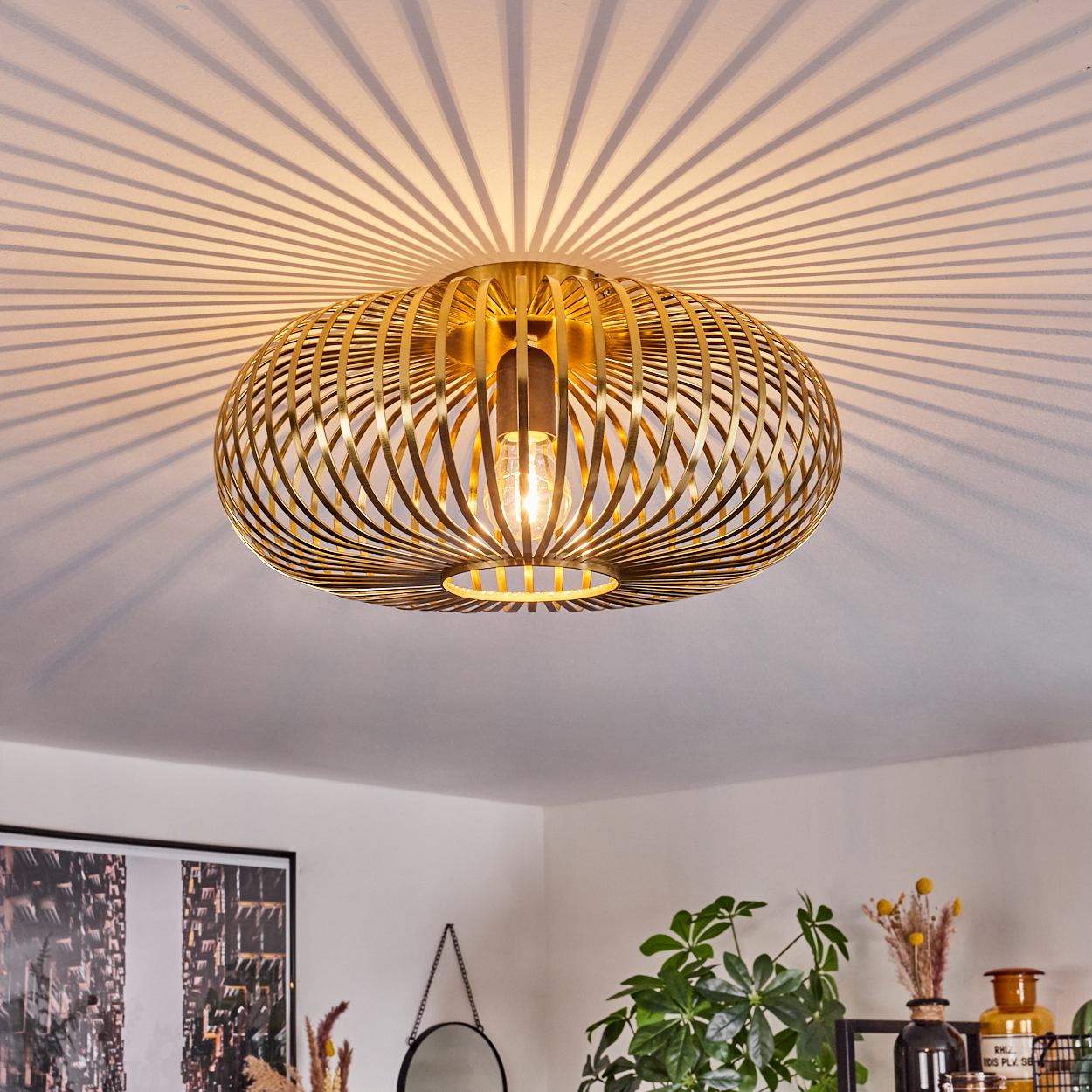 Eine goldene, käfigartige Deckenleuchte wirft einen radialen Schatten auf die weiße Decke. Unten Andeutungen von Dekor: Kunst, Spiegel und Pflanzen.