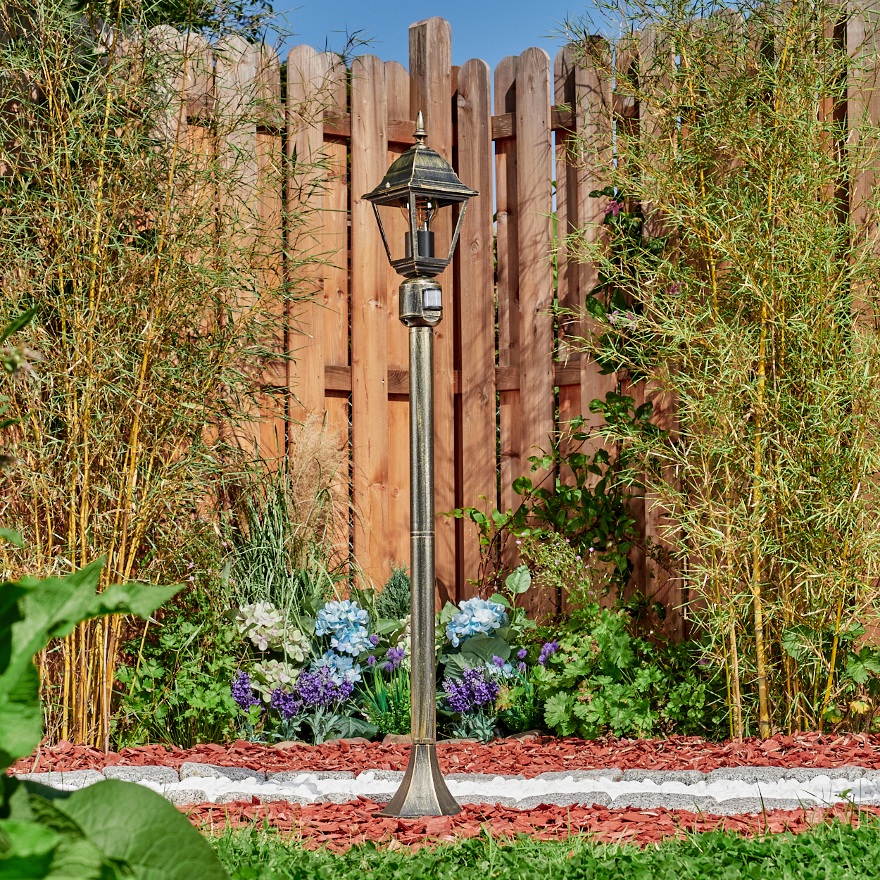 Eine bronzefarbene Straßenlaterne steht in einem Garten mit Blumen und Bambus. Im Hintergrund befindet sich ein Holzzaun unter blauem Himmel.