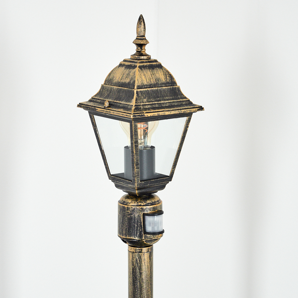 Eine schwarze und goldene Außenlaterne mit einem Gehäuse aus klarem Glas. Die Lampe hat eine spitze Spitze und einen Bewegungssensor.