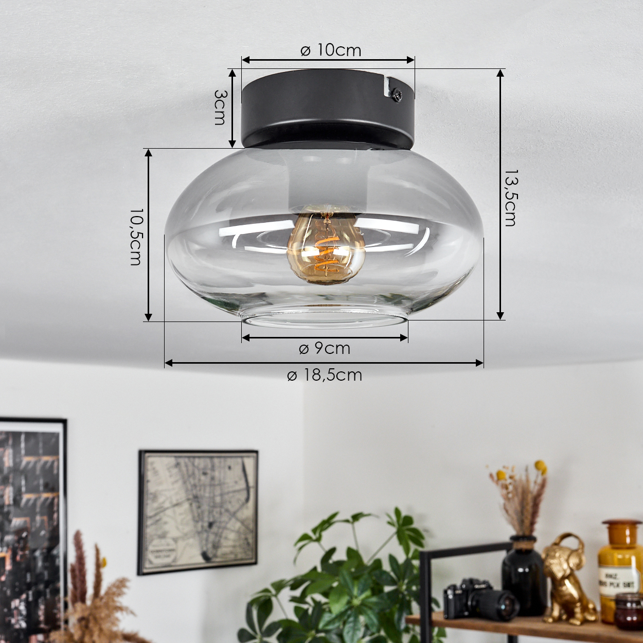Eine Deckenlampe aus Glas mit Maßangaben. Darunter ein Regal mit Deko, Kamera, goldenem Hund und Wandkarte.