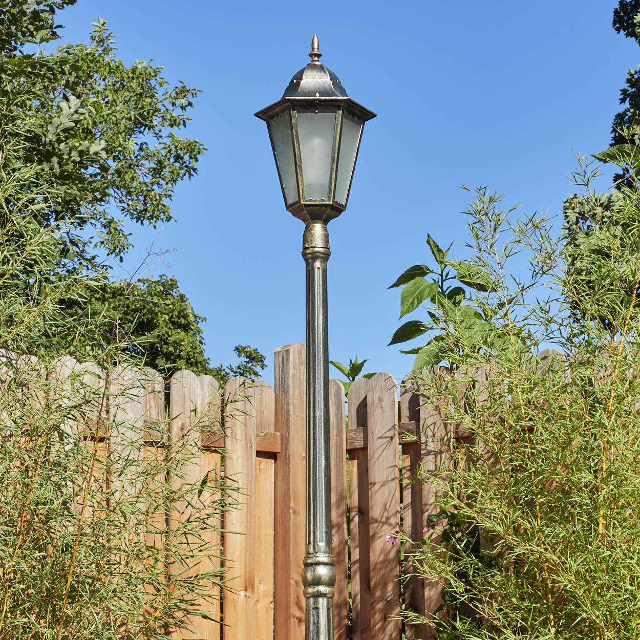 Eine dekorative Laterne in einem Garten. Dahinter ein Holzzaun und grüne Pflanzen mit blauem Himmel.