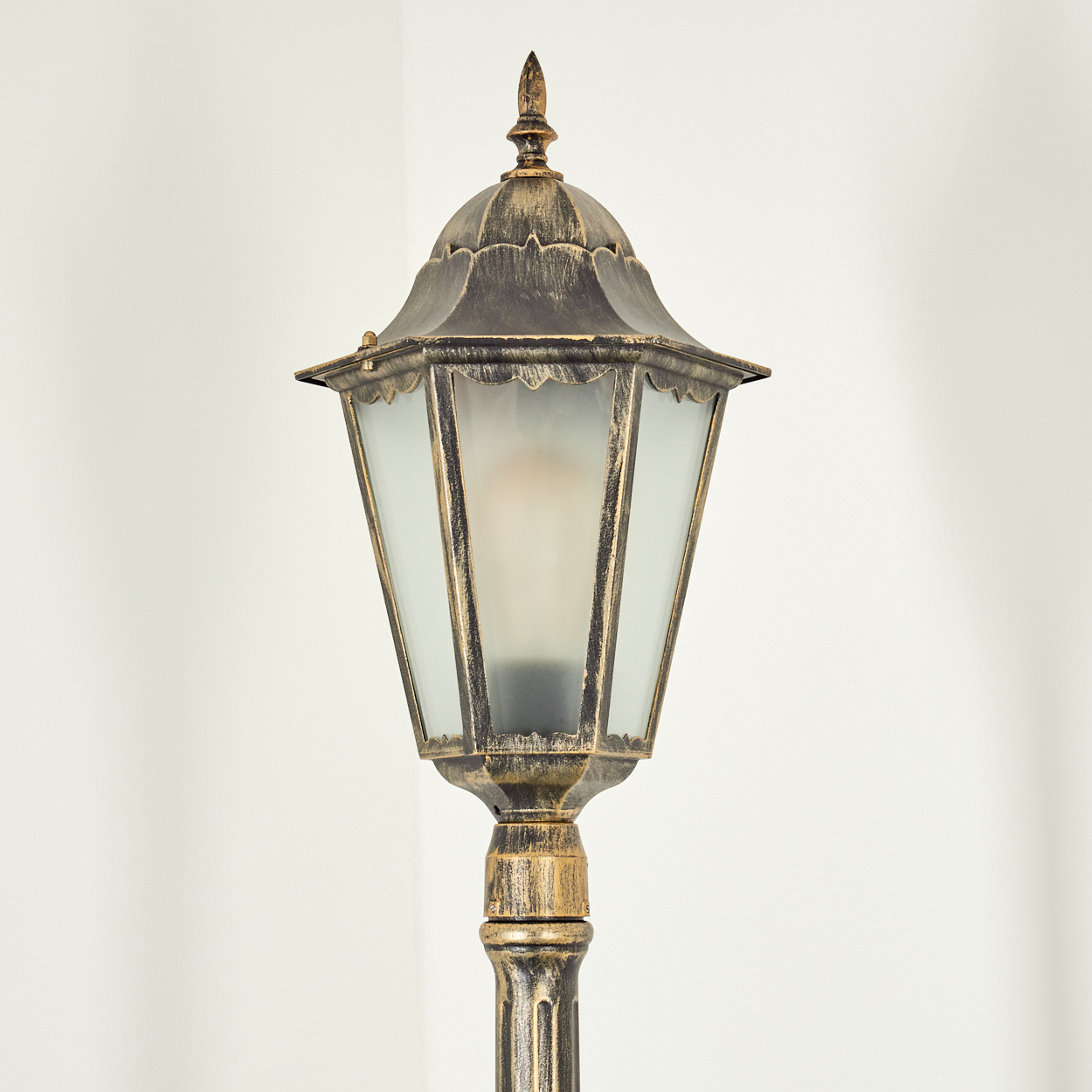 Eine verzierte, antike Außenlampe mit Bronzefinish steht vor einer weißen Wand. Die Lampe hat Glasscheiben.