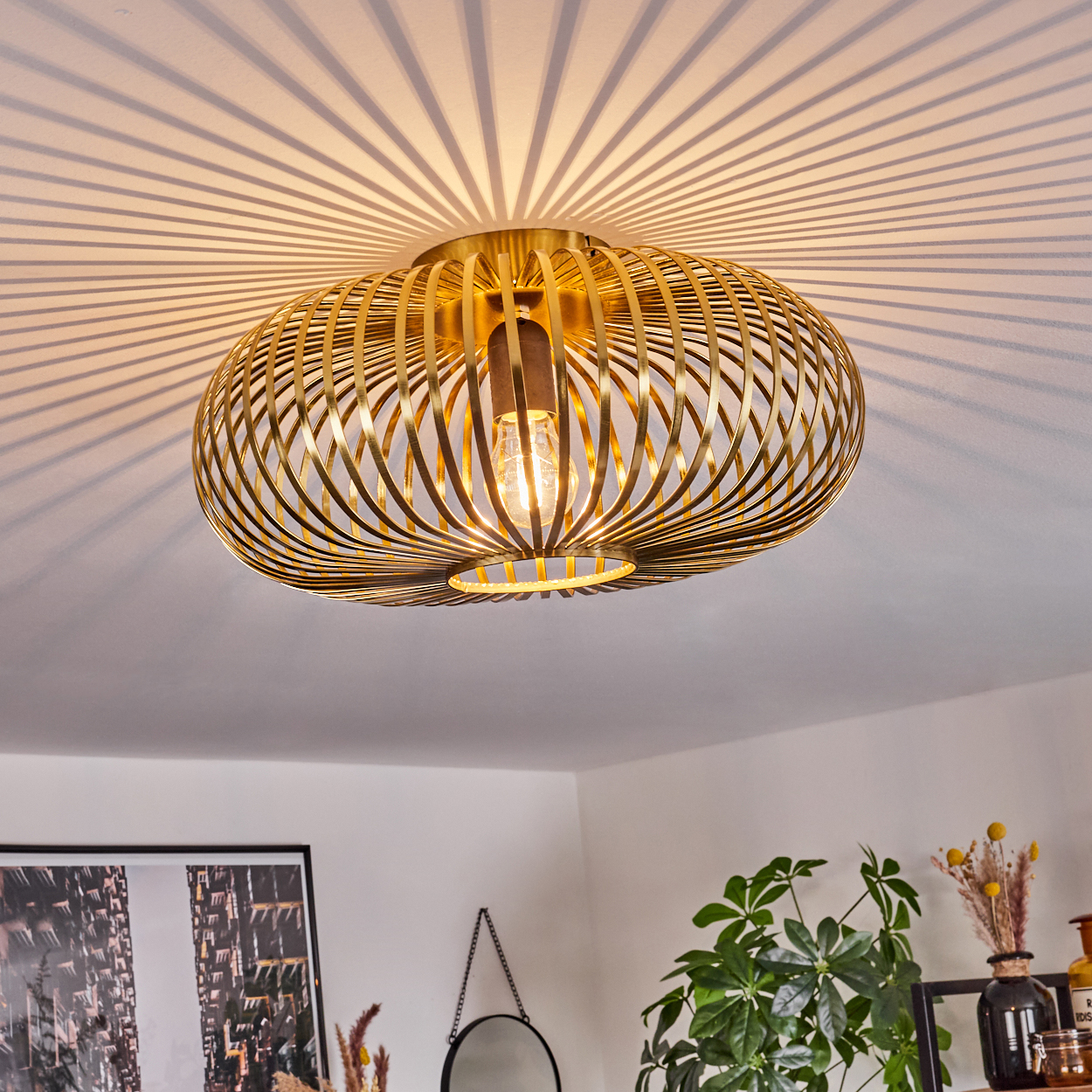 Eine goldene, käfigartige Deckenlampe wirft radiale Schatten. Darunter treffen eine weiße Decke und Wände aufeinander. Dekorationen und Pflanzen sind sichtbar.