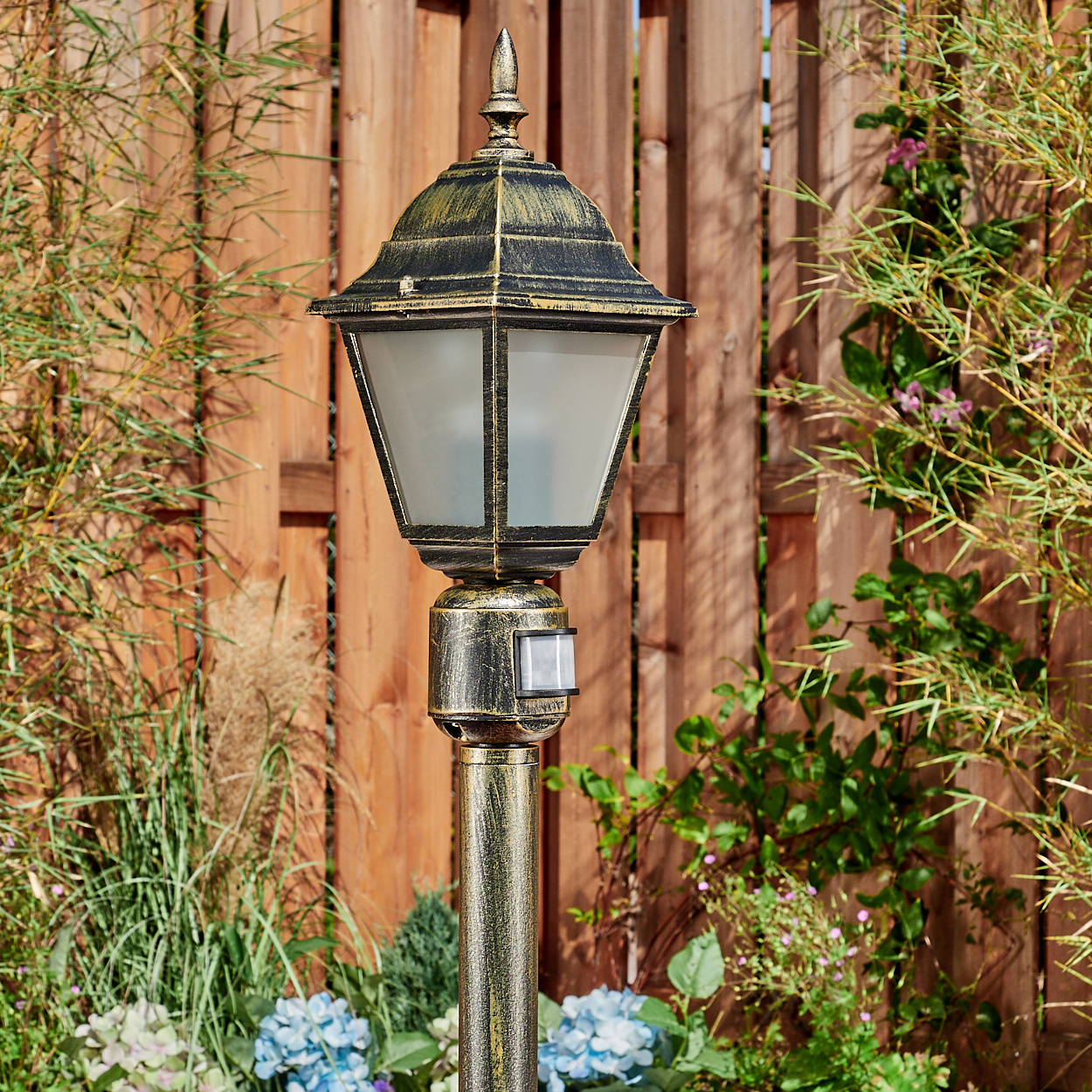 Eine verzierte, goldfarbene Außenlaterne mit einem Schirm aus Milchglas steht in einem Garten. Im Hintergrund ist ein Holzzaun zu sehen.