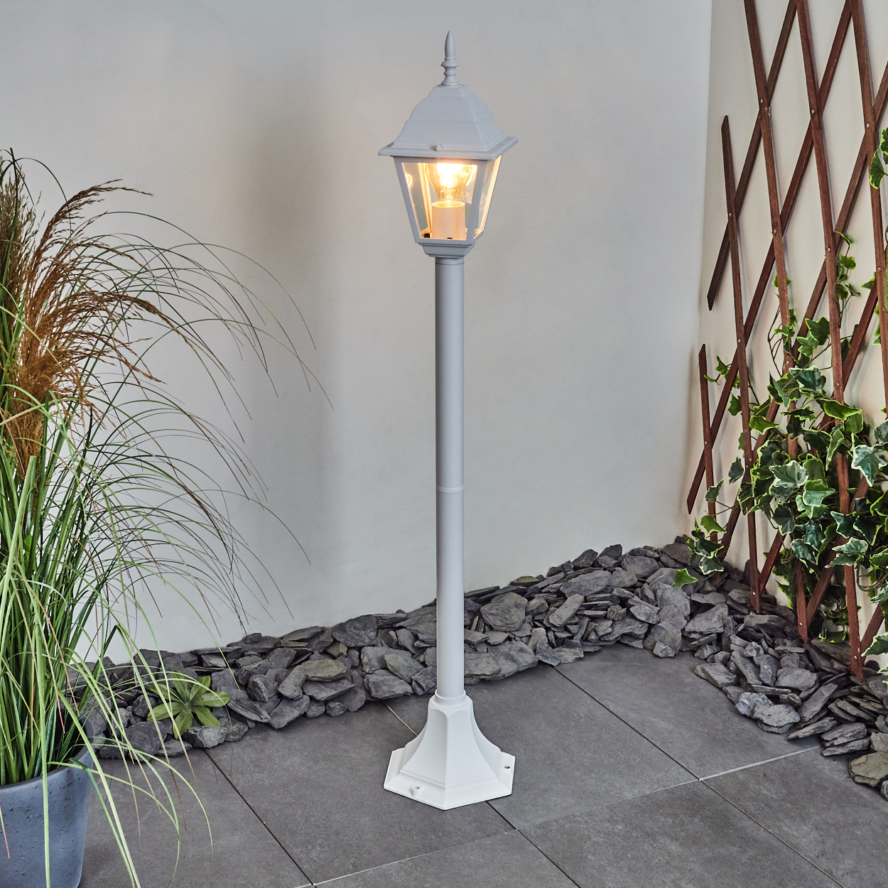 Eine weiße Laterne mit leuchtender Glühbirne steht auf einer grauen Fliesen-Terrasse. Pflanzen und Steine säumen die Lampe. Ein Spalier und Efeu sind sichtbar.