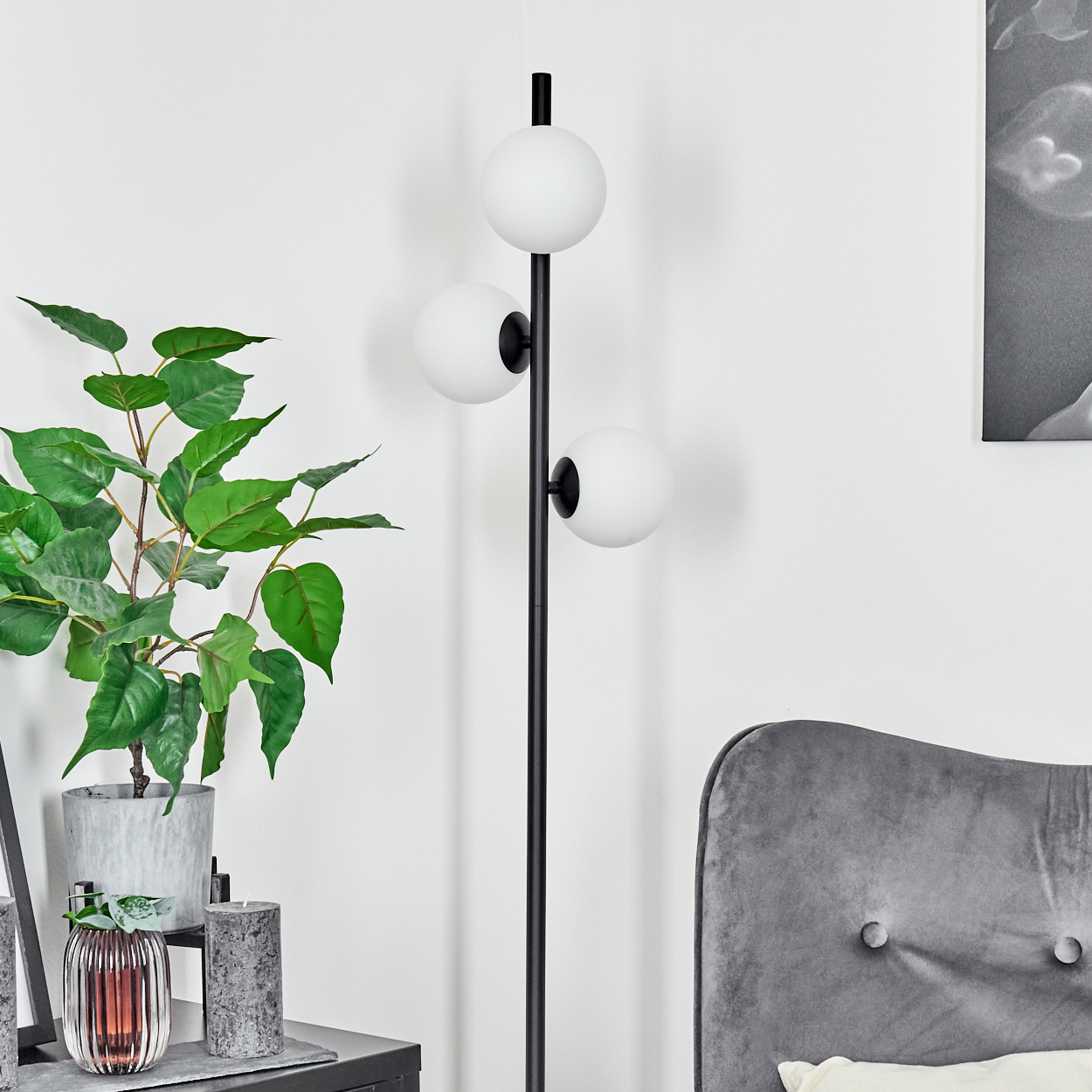 Eine moderne Innenszene mit einer schwarzen Stehlampe mit drei weißen Kugelleuchten, einer Topfpflanze und einem grauen Samtstuhl.