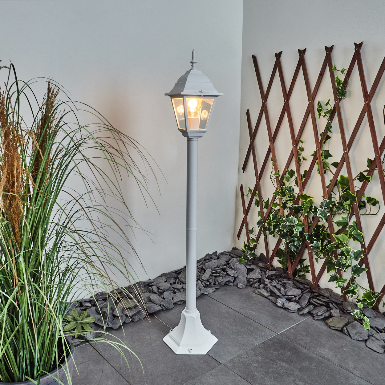 Eine graue Laterne mit einer leuchtenden Glühbirne steht in einem Garten. Rechts sind grüne Pflanzen und ein Holzgitter mit Efeu.